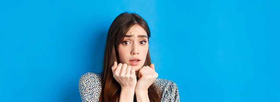 Close-up of scared timid girl looking afraid, shaking from fear, press hands to body and staring frightened, standing on blue background.