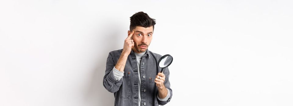 Confused guy found something down with magnifying glass, look puzzled at camera, standing against white background.