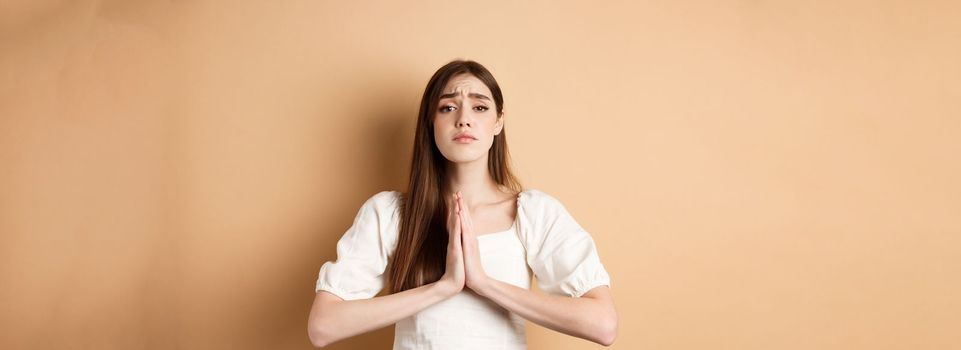 Please help. Desperate young woman begging you, holding hands in pleading gesture and looking sad at camera, asking for favour, beige background.