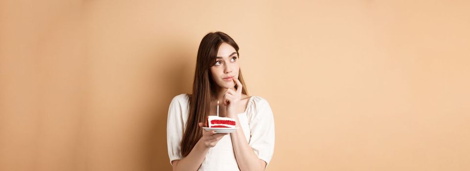 Dreamy birthday girl look aside and thinking of wish, blowing candle on cake, standing on beige background.