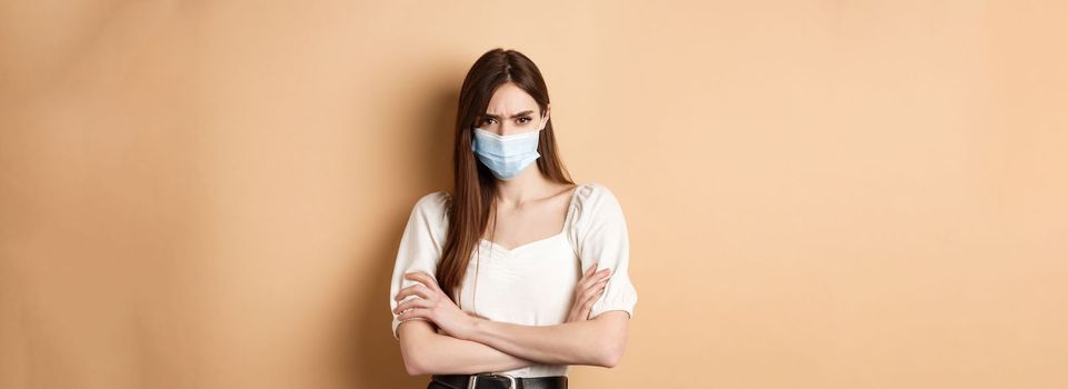 Covid-19 and lifestyle concept. Angry woman look with despise, wearing medical mask, cross arms on chest and stare with condemn, standing on beige background.