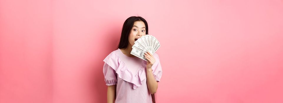 Cute asian woman showing dollar bills and smiling amazed. Rich woman waiving with money, standing against pink background.