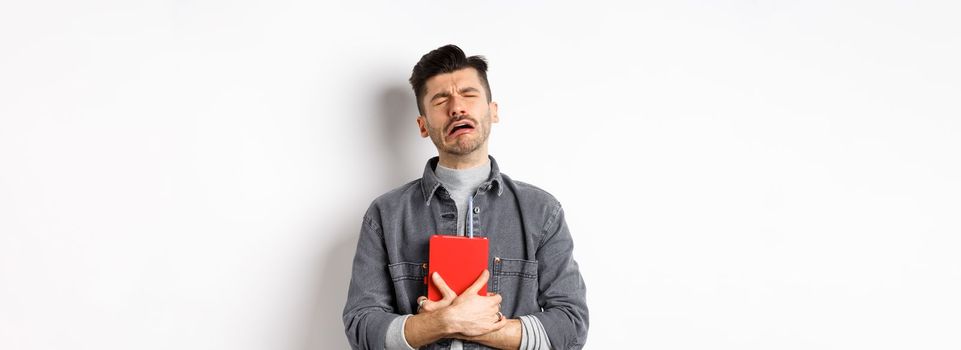 Sad cryig man holding red diary and sobbing, miserable guy carry journal with him, standing against white background.