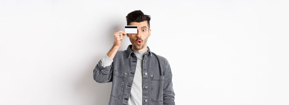 Excited funny man hold plastic credit card on eye and say wow, checking out special deal, standing on white background.