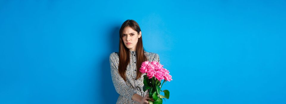 Valentines day concept. Displeased young woman rejecting flowers gift, frowning and grimacing, refusing take roses, standing on blue background.