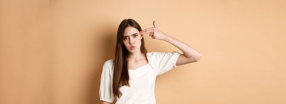 Just kill me. Annoyed and distressed young woman making finger gun sign on ver head and frowning bothered, standing pissed-off on beige background.