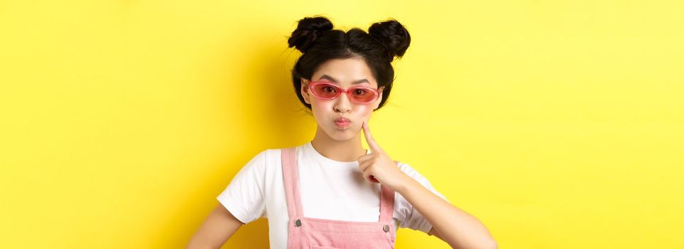 Stylish glam girl with sunglasses, pouting and poking cheek, standing silly on yellow background.