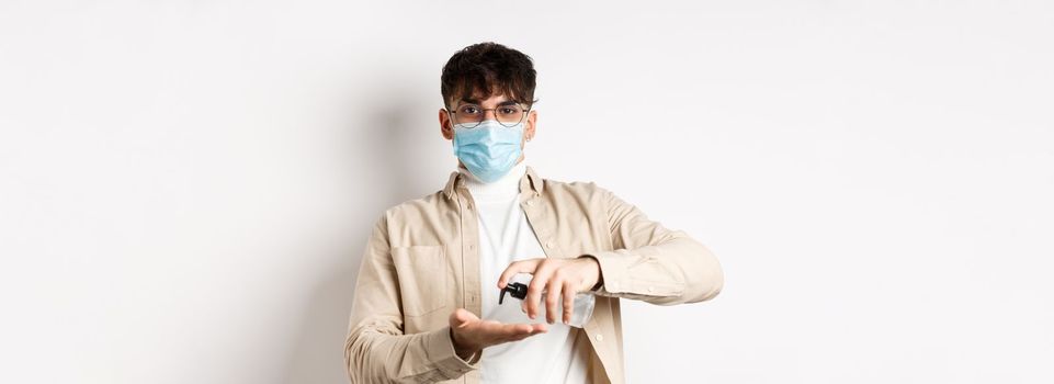 Health, covid and quarantine concept. Young hispanic guy in glasses and face mask using hand sanitizer, apply antiseptic and look at camera, standing on white background.