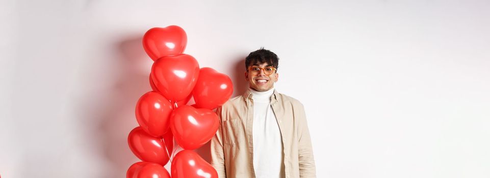 Valentines day. Excited young man smiling, looking hopeful, standing near big red hearts balloons, waiting for true love on lovers date, white background.