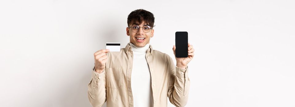 Online shopping. Surprised and happy young man showing credit card and mobile phone screen, standing on white background.