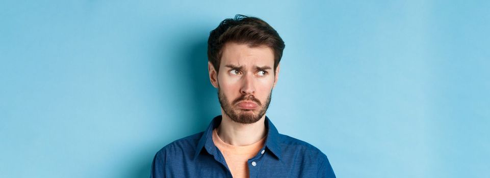 Miserable and disappointed guy acting childish, sulking and looking left at empty space with offended face, standing on blue background.