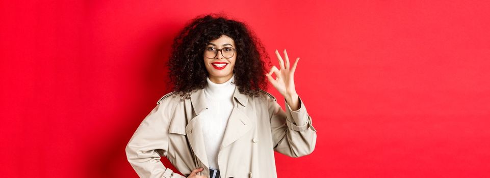 Portrait of confident trendy woman in glasses and trench coat, showing okay gesture to approve or agree with you, say yes, standing on red background.