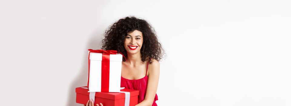 Beautiful birthday girl with curly hair, holding bday gifts and smiling happy, celebrating, standing against white background.