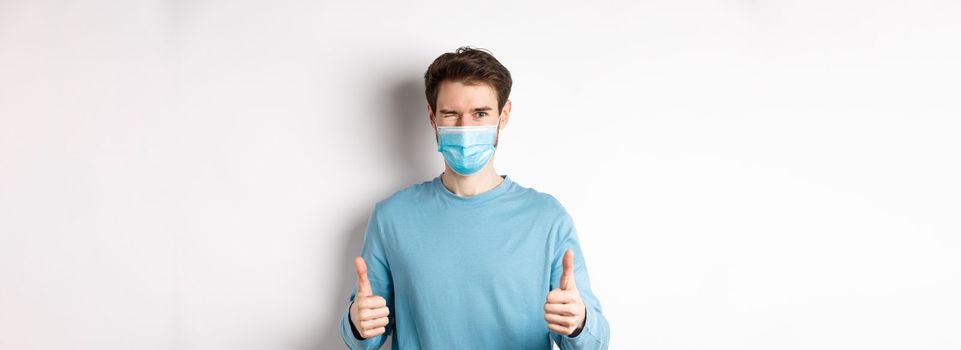 Covid-19, pandemic and social distancing concept. Happy young man in medical mask winking, showing thumbs up in approval, recommending product, white background.