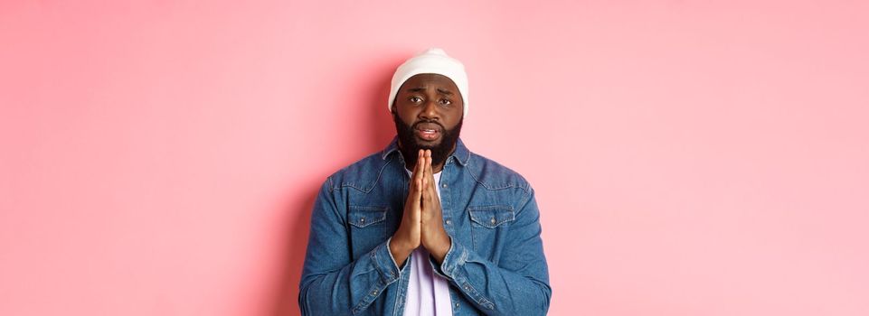 Upset african-american man asking for help, holding hands in pray, staring at camera sad and pleading, standing over pink background.