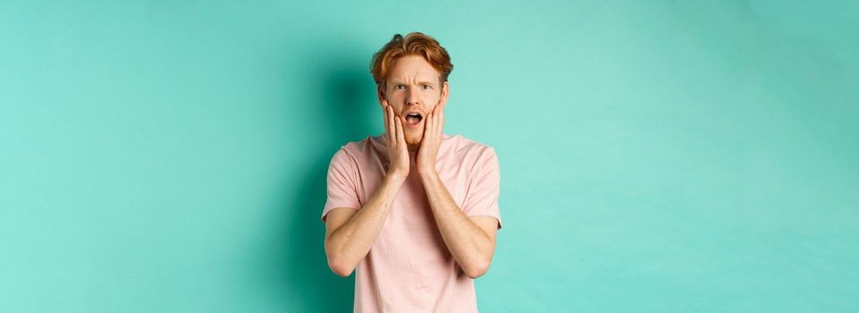 Shocked and concerned young man with red hair, staring at camera worried and touching face, standing in t-shirt against turquoise background.