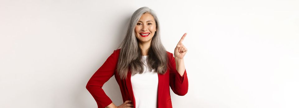 Happy senior office lady pointing finger at upper left corner, showing company logo and smiling at camera, standing over white background. Copy space