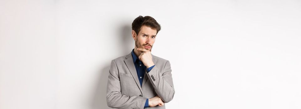 Hesitant young businessman in suit looking skeptical at camera, thinking and making decision, standing against white background.