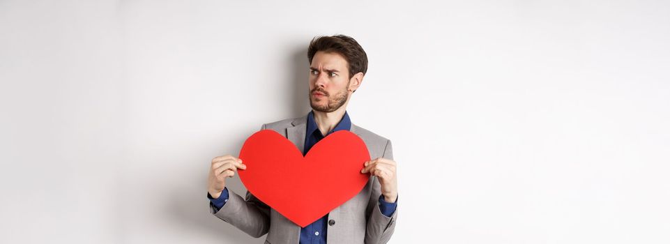 Confused man in suit holding big red heart and looking right, searching for love on Valentines day, standing over white background.