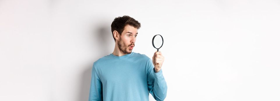 Man got surprised while looking through magnifying glass, saying wow at awesome product, standing over white background.
