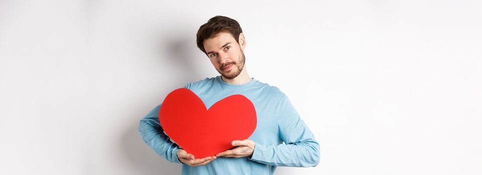 Romantic boyfriend making Valentines day surprise, holding big red heart cutout on chest and smiling with love, looking tender at camera, standing over white background.