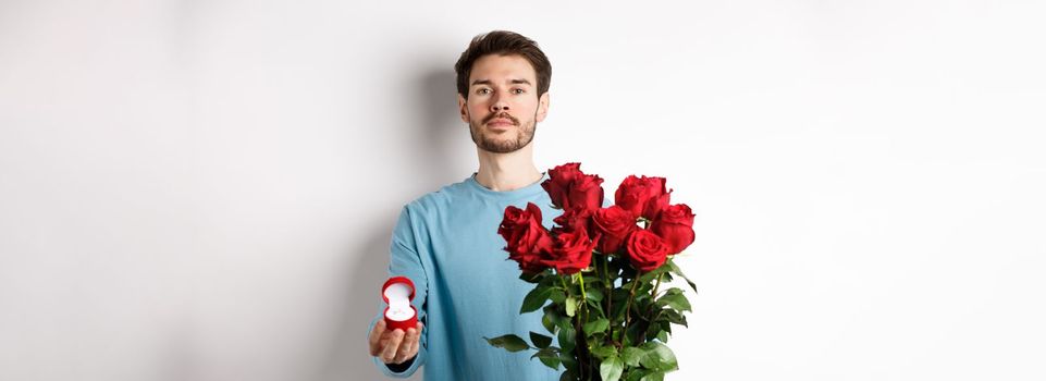 Valentines and relationship. Romantic man boyfriend holding red roses and showing engagement ring, making a proposal on lovers day, standing over white background.