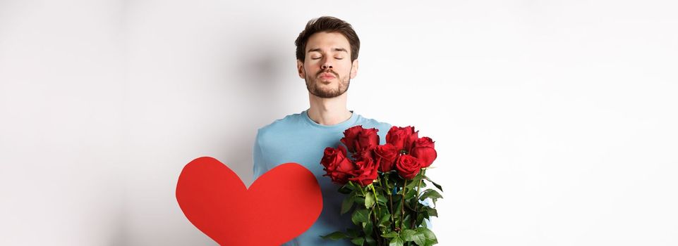 Romantic boyfriend waiting for kiss, holding bouquet of roses flowers and big red heart on Valentines day, love in air, standing over white background.