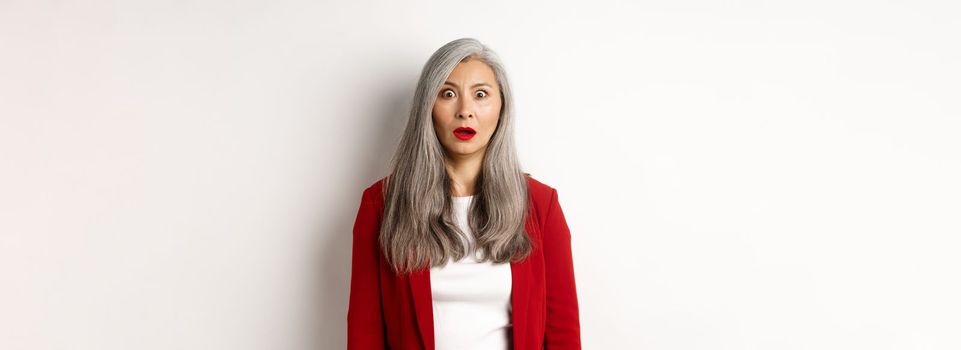 Business concept. Shocked senior female employer staring at camera speechless, standing in red blazer and makeup over white background.