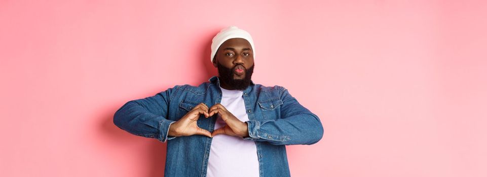 Happy african-american man showing heart sign, I love you gesture, pucker lips for kiss while standing over pink background.