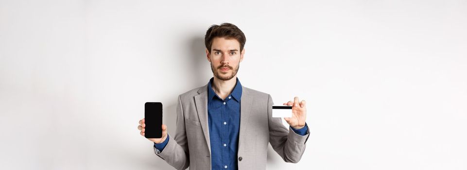 Online shopping. Serious bearded man in business suit showing plastic credit card with empty smartphone screen, standing against white background.