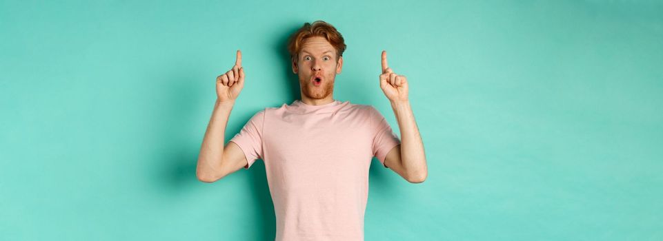 Impressed handsome guy with red hair pointing fingers up, demonstrate promo offer, standing in t-shirt over mint background.