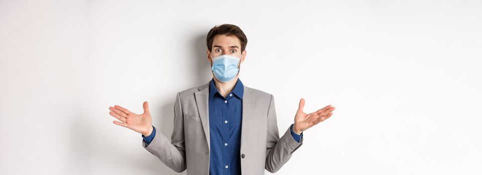 Health, pandemic and business concept. Confused businessman in medical mask and suit spread hands sideways and look puzzled, cant understand, white background.