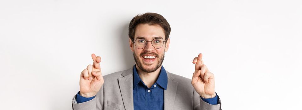 Close-up of hopeful male entrepreneur in glasses and suit cross fingers for good luck, smiling and making wish, praying for good results, white background.