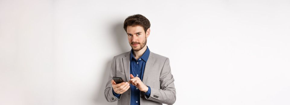 Handsome bearded male model in suit using mobile phone, smiling pleased at camera, white background.