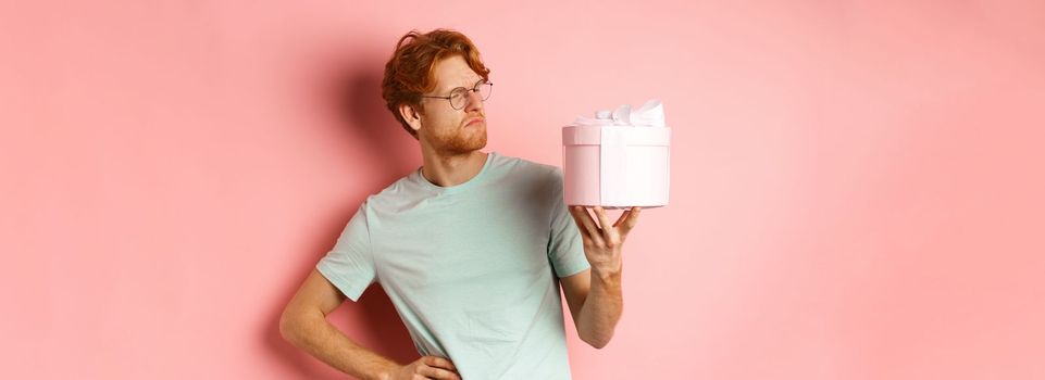 Love and holidays concept. Intrigued redhead guy looking puzzled at gift box, dont know what inside, standing over pink background.