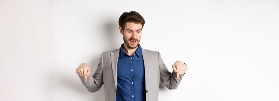 Excited guy in suit smiling and pointing fingers down, looking at special deal with amused face, standing on white background.