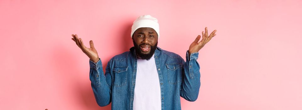 Confused and bothered Black man complaining, shrugging and raising hands up, staring with disappointment, standing over pink background.