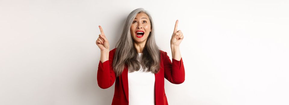 Happy asian lady in red blazer and makeup, looking and pointing fingers up, showing special offer, standing over white background.