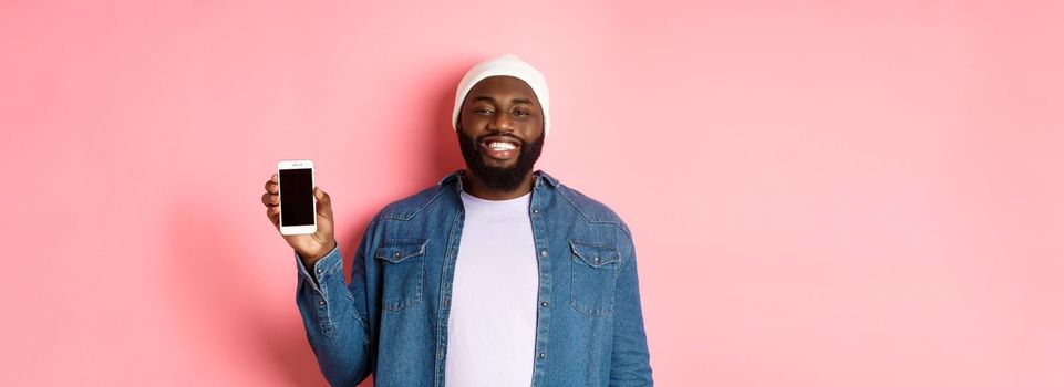 Handsome hipster guy in beanie and denim shirt smiling, showing mobile phone screen with happy face, introduce application, standing over pink background.