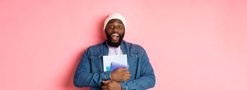 Image of adult african-american man holding notebooks and smiling, studying at courses, standing over pink background.