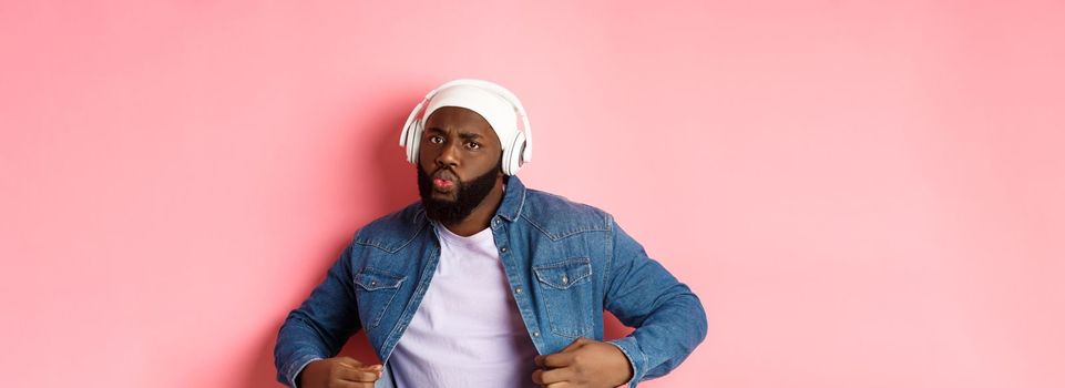 Cool and sassy Black guy dancing, listening to music in headphones, looking confident, standing over pink background. Copy space