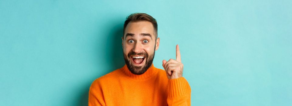 Close-up of handsome man having an idea, raising finger up and smiling excited, standing in orange sweater.