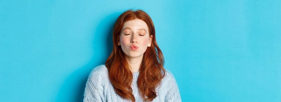Close-up of young woman with red wavy hair standing with eyes closed and puckered lips, waiting for kiss against blue background.