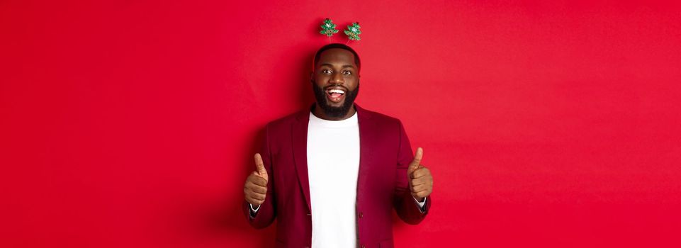 Merry Christmas. Happy african american man celebarting New Year, wearing funny party headband and showing thumb up, like and praise something, standing over red backgrond.