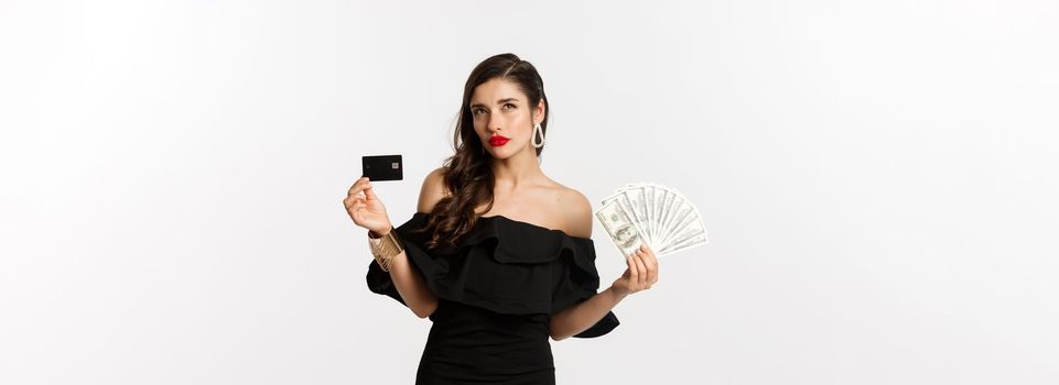 Fashion and shopping concept. Thoughtful woman holding credit card and dollars, thinking and looking up, white background.