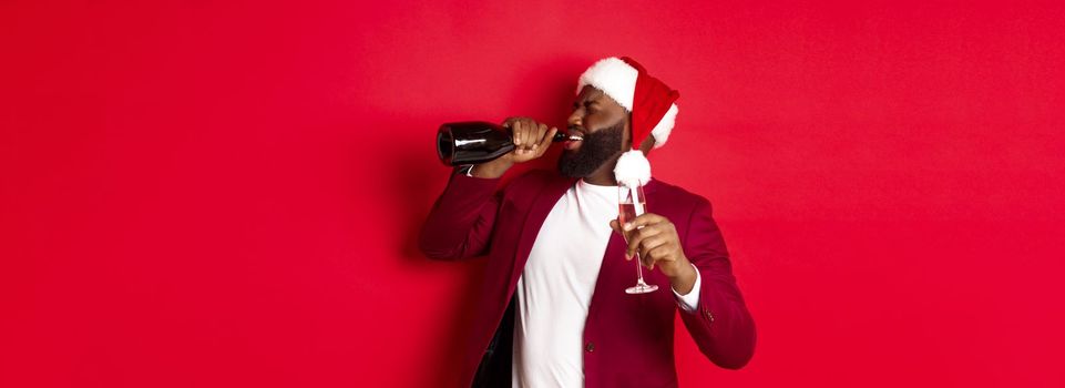 Christmas, party and holidays concept. Image of young Black man in santa hat drinking champagne from bottle, getting drunk on New Year celebration, standing over red background.