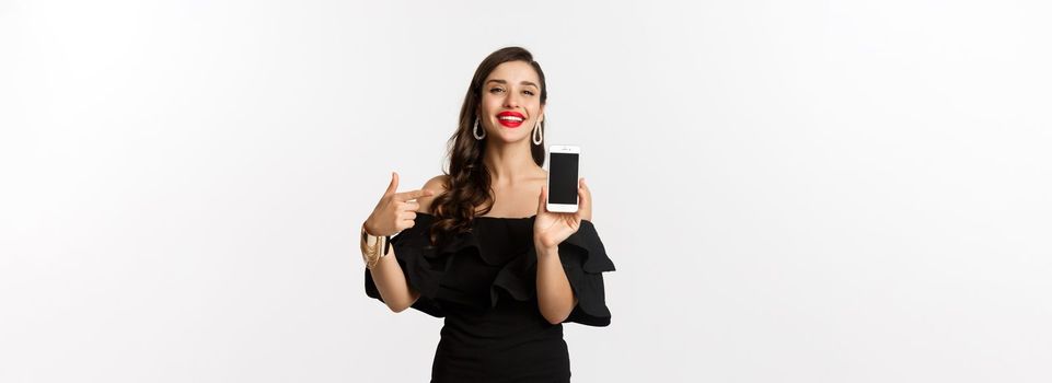 Online shopping concept. Fashionable woman in black dress pointing finger at smartphone screen, showing application, standing over white background.