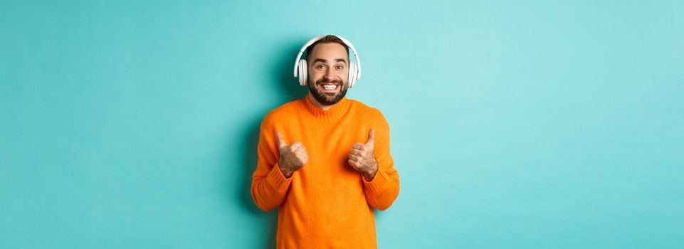 Amazed adult man listening music in headphones, looking at camera impressed and showing thumbs-up in approval, recommending, standing over turquoise background.