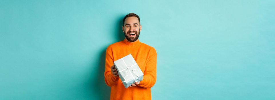 Holidays and celebration concept. Cheerful bearded man giving present, holding gift and smiling, congratulating you, standing over turquoise background.