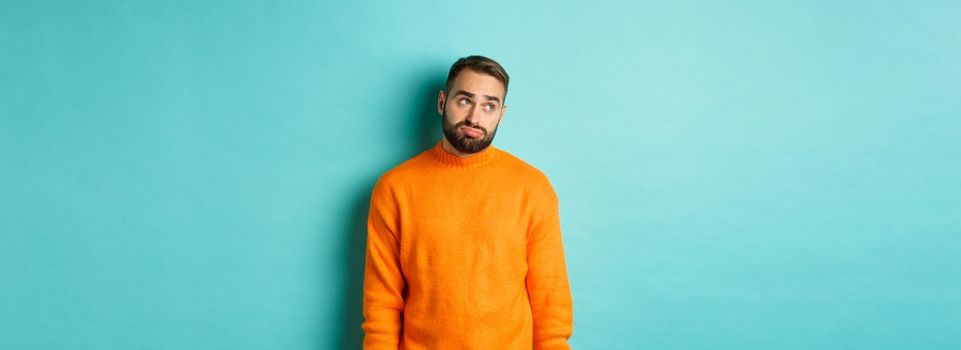 Sad and gloomy man sulking, looking bored at upper left corner, standing in orange sweater against light blue background.